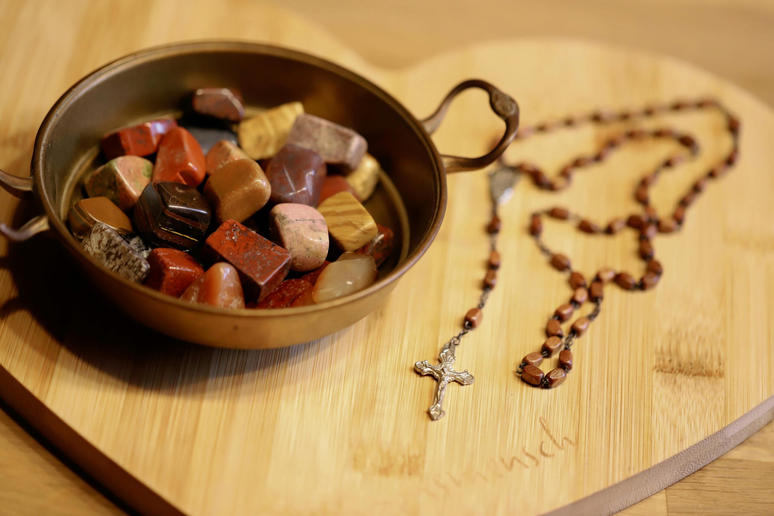 Colorful healing stones in bowl alongside rosary on wooden board symbolizing spirituality and tranquility.